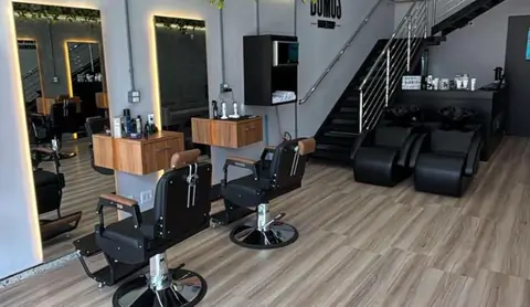 Cadeiras de barbeiro e bancadas de madeira da Domus Barbershop, com espelhos iluminados e a escada ao fundo